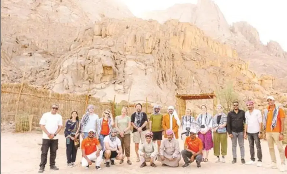 A group of visitors and local hosts stand together on sand in front of a bare desert mountain, beside a reed fence and a wooden gateway. Published with the Al-Nahar report on the Bedouin heritage centre near Hurghada.