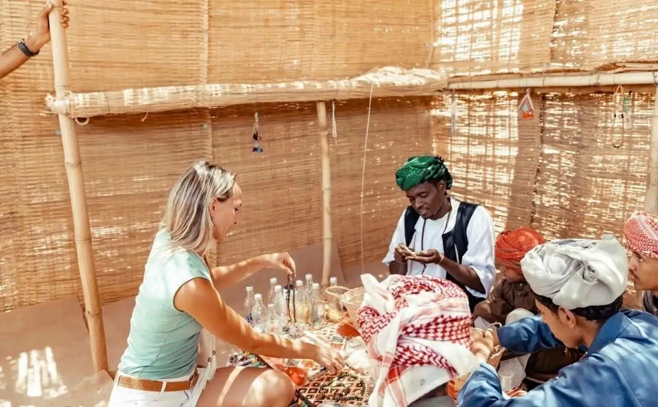A visitor sits with Bedouin hosts on a rug inside a reed-walled desert shelter, handling beadwork and other crafts. Photograph published with the report on the Bedouin heritage tourism project near Hurghada.