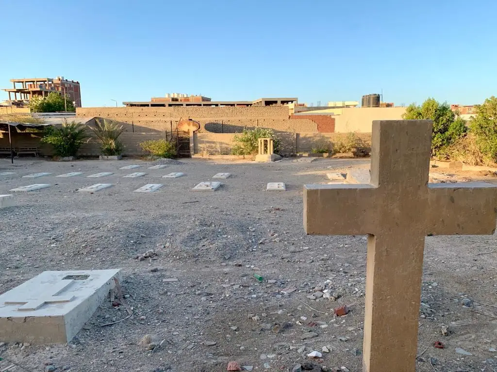 A walled cemetery with flat stone grave slabs on open ground and a large stone cross in the foreground, town buildings beyond the wall. Published by Youm7 with its feature on El Quseir's Italian phosphate era; the location shown is not independently confirmed.