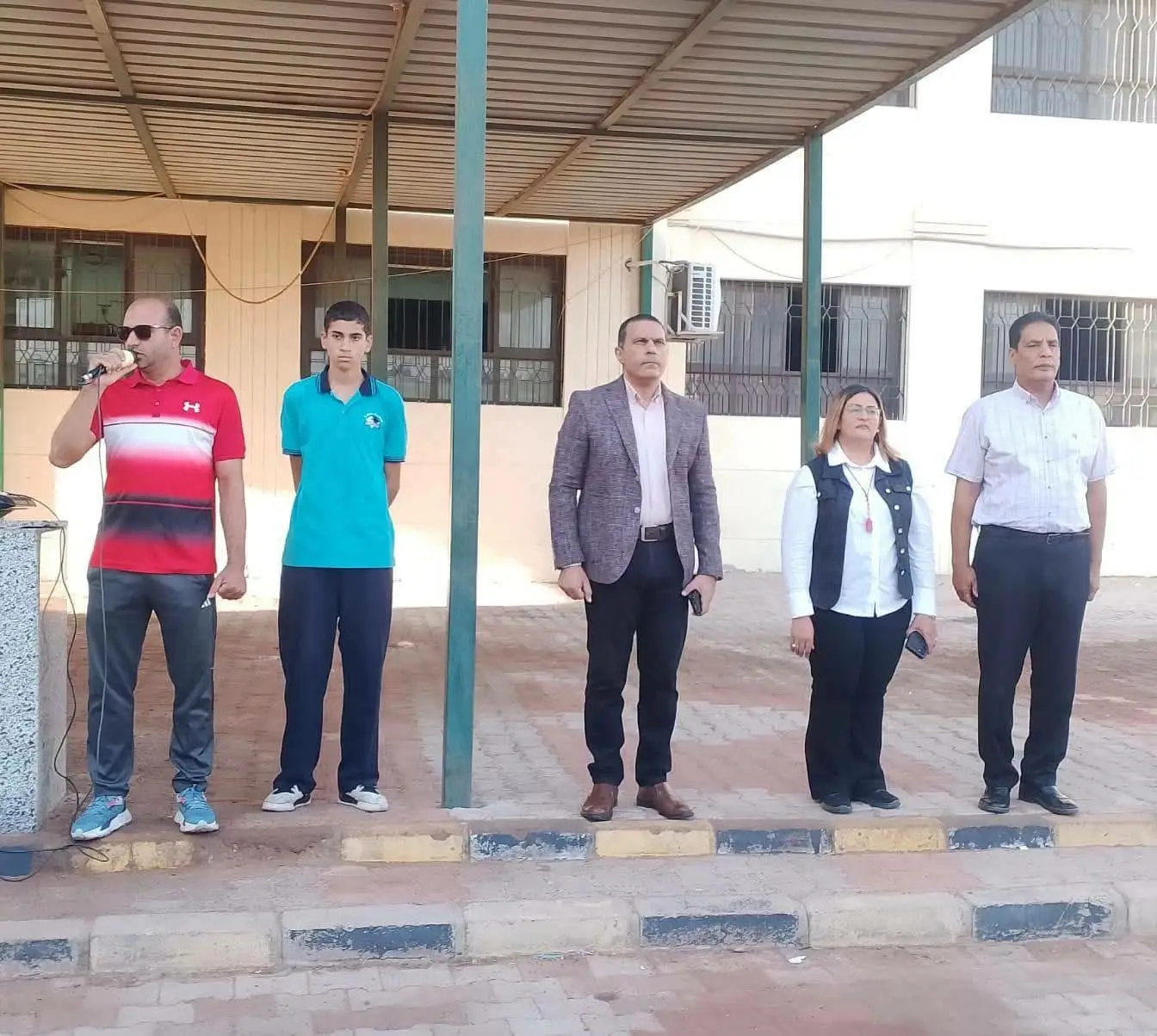 Five people, among them a pupil in school uniform, stand in a school yard under a shaded walkway during a morning assembly, one man speaking into a microphone. Photograph published by Al-Masry Al-Youm with its report on the inspection order; the individuals are not identified in the report.