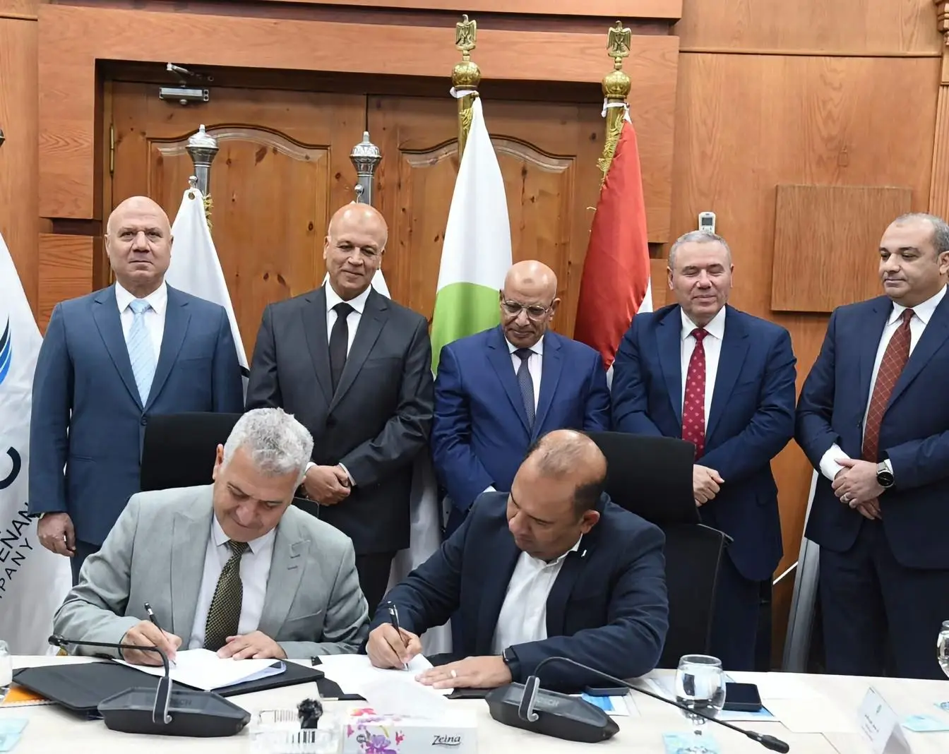 Two men sign documents at a conference table while five officials stand behind them; a banner for the Egyptian Maintenance Company and Egyptian flags are in the background. The publisher ran this photograph with its report on the desalination contracts.